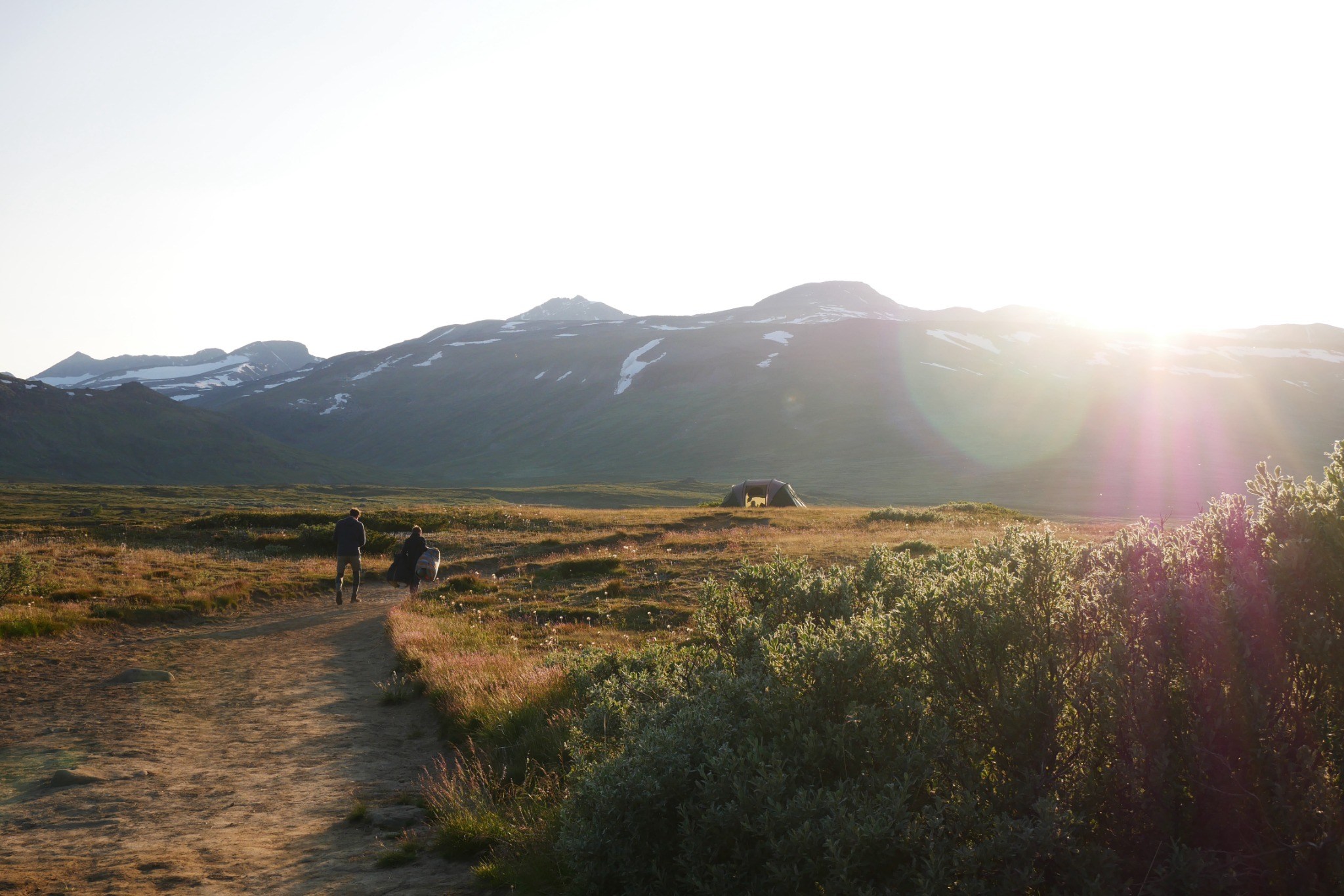 Two people walking along a path through a mountain landscape in evening light